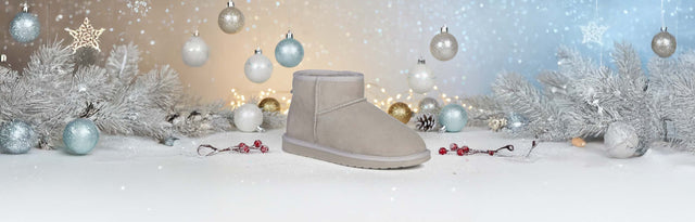 Stivaletti invernali EMU Australia in suede grigio con fodera in pelliccia, appoggiati su sfondo verde petrolio accanto a un morbido tessuto in lana.