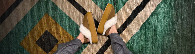 Stivaletti invernali EMU Australia in suede grigio con fodera in pelliccia, appoggiati su sfondo verde petrolio accanto a un morbido tessuto in lana.