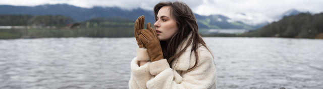 Donna con guanti EMU Australia in pelle scamosciata marrone con interno shearling, abbinate a cappotto invernale chiaro, in riva a un lago di montagna.