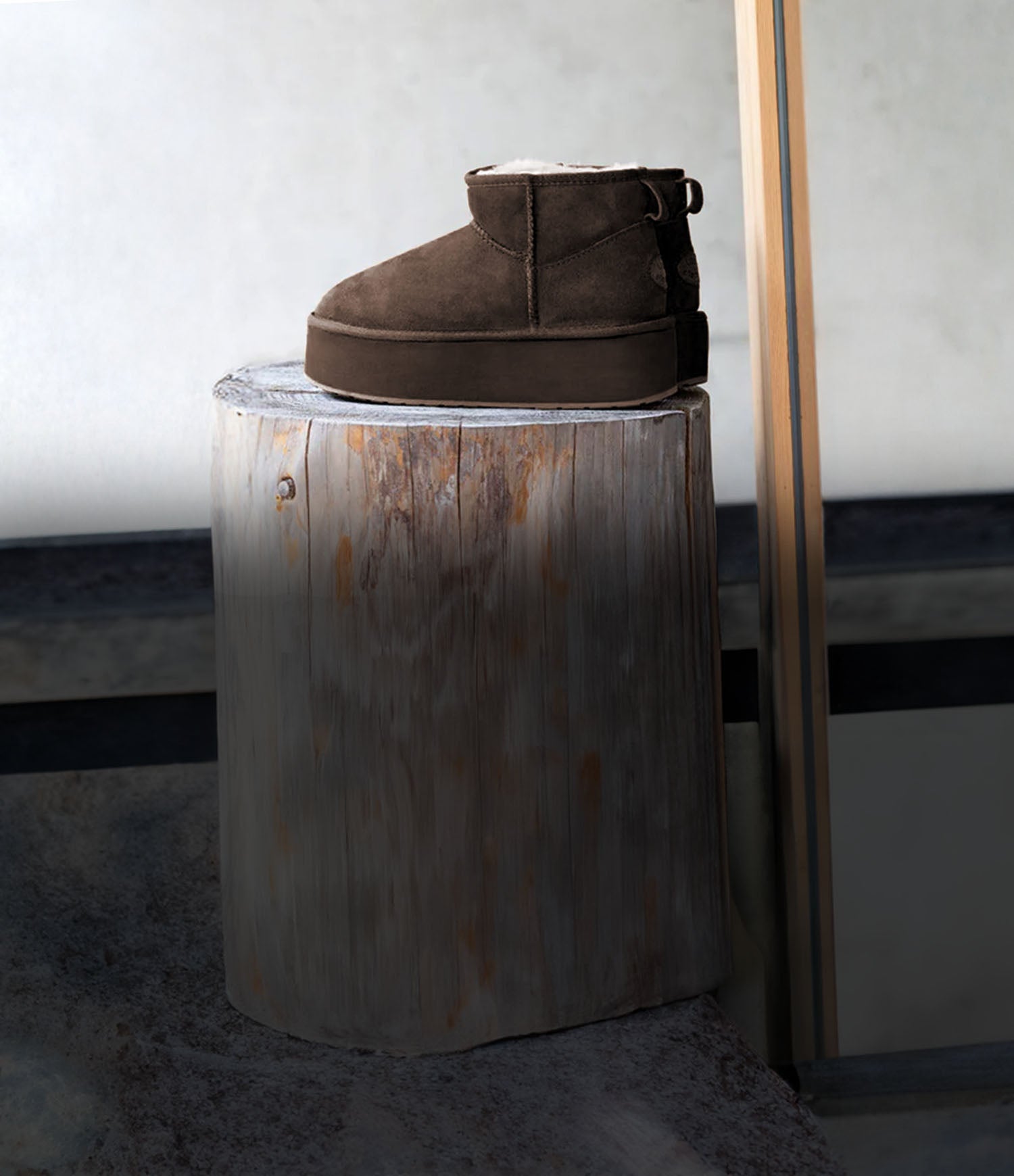 Stivaletto in pelle scamosciata marrone EMU Australia posizionato su un tronco di legno, con interno in lana visibile, in un set minimal da studio.