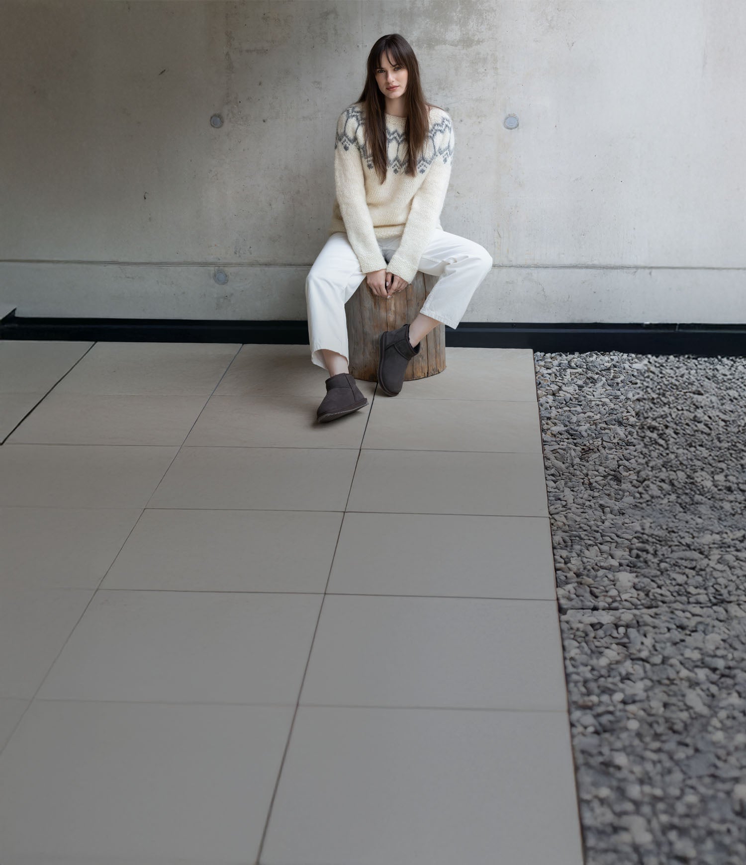 Modella seduta in un ambiente minimal che indossa stivaletti invernali EMU Australia grigi, abbinati a maglione chiaro e pantaloni bianchi.