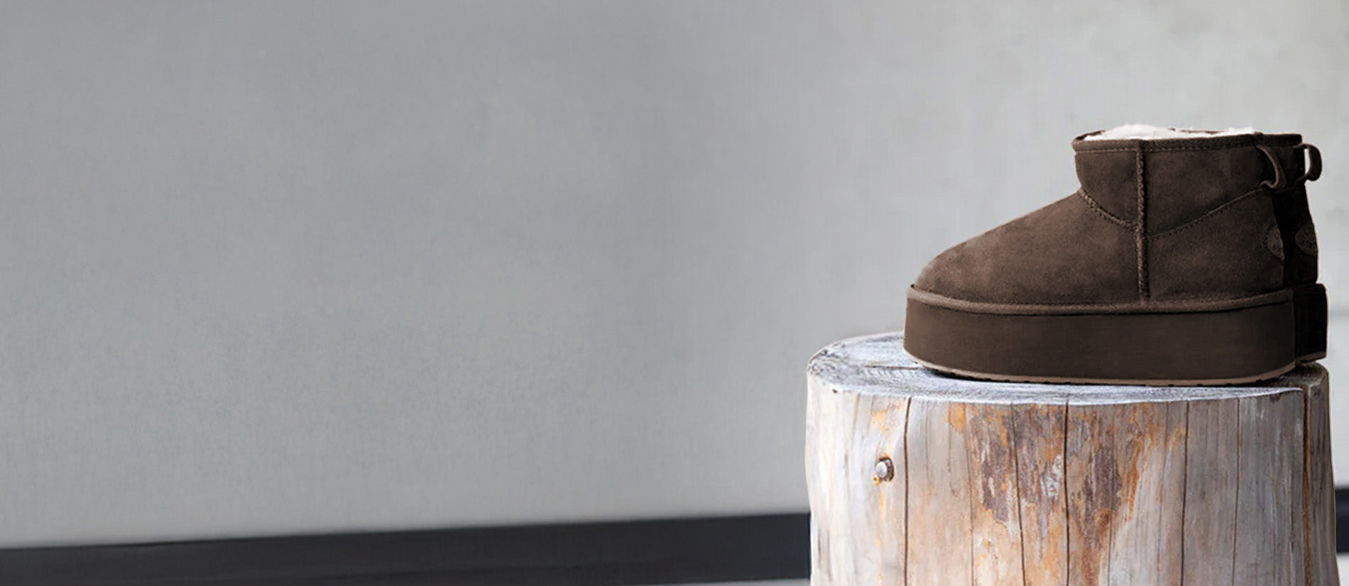 Stivaletto in pelle scamosciata marrone EMU Australia posizionato su un tronco di legno, con interno in lana visibile, in un set minimal da studio.