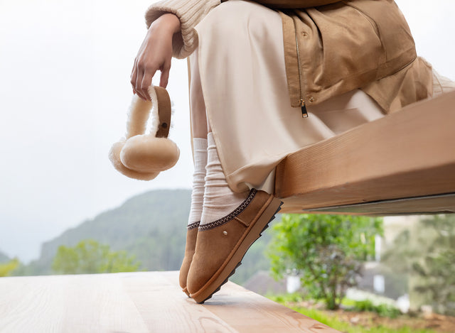 Piedi di una donna che indossa ciabatte invernali EMU Australia Sharky Scuff color cognac in pelle scamosciata, con cuffie auricolari in shearling in mano, in ambientazione outdoor.