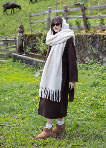 Donna con cappotto lungo marrone e maxi sciarpa bianca, indossa stivaletti EMU Australia in shearling marrone, collezione invernale