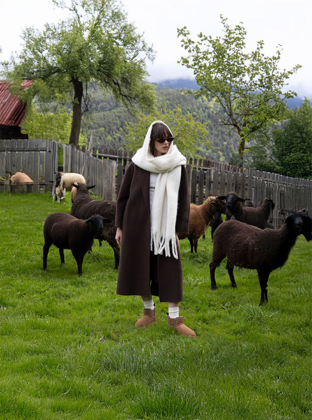 Donna con stivaletti shearling EMU Australia e cappotto marrone, tra pecore al pascolo in montagna, collezione The EMU Way