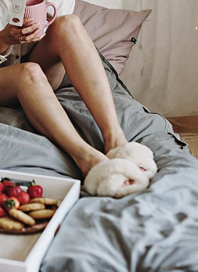 Donna rilassata sul letto con pantofole morbide in tessuto teddy color panna, atmosfera cozy e домашняя, ideale per comfort indoor e momenti di relax.