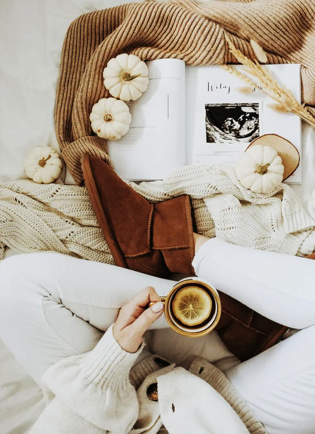 Donna in relax con stivaletti in pelle scamosciata marrone e interno in shearling, circondata da coperte e dettagli autunnali in un’atmosfera calda e accogliente.