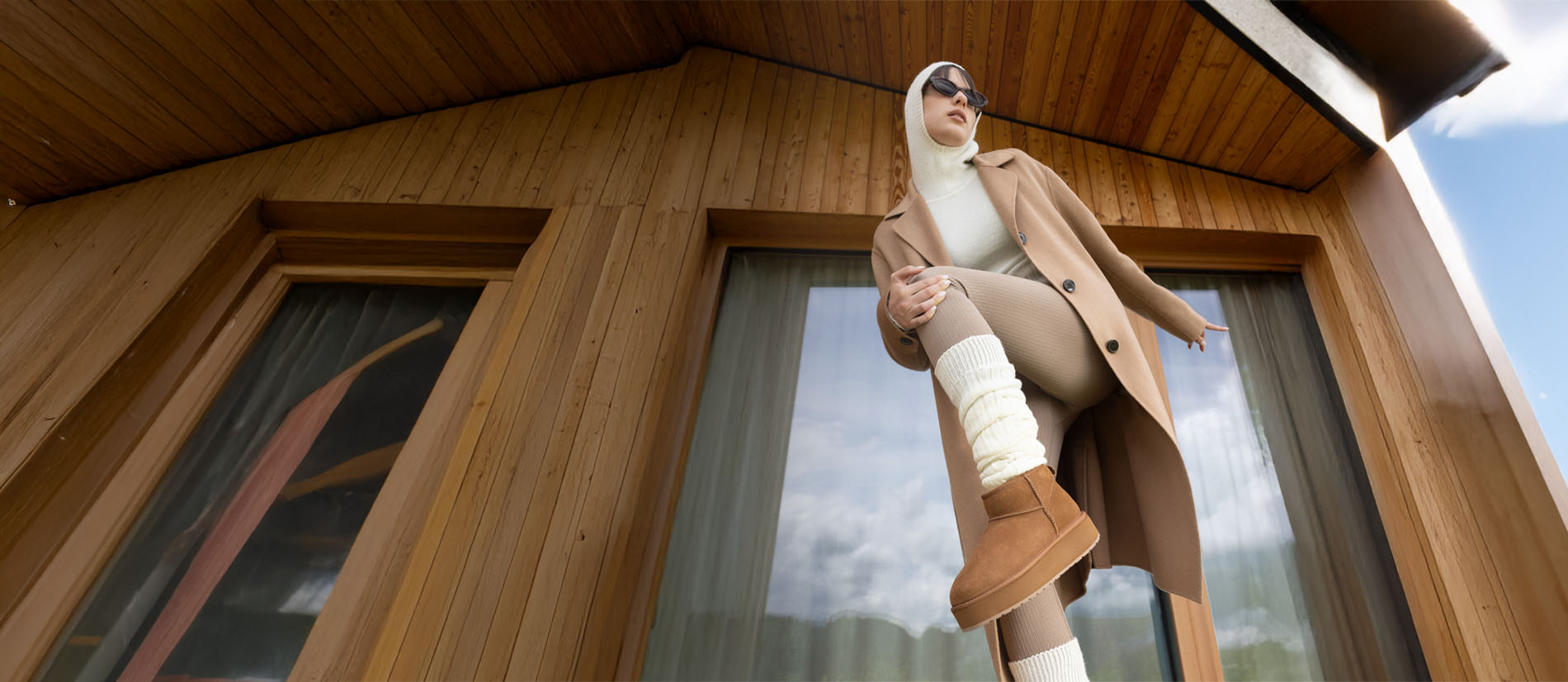 Donna con outfit invernale beige che indossa stivaletti EMU Australia marroni all’esterno di una casa in legno, in stile lifestyle outdoor.