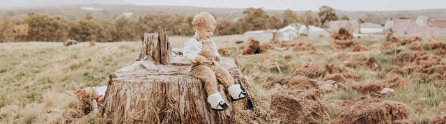 bambino piccolo seduto su un tronco in mezzo alla campagna che indossa stivaletti bianchi e neri con pelo emu australia shaun the sheep